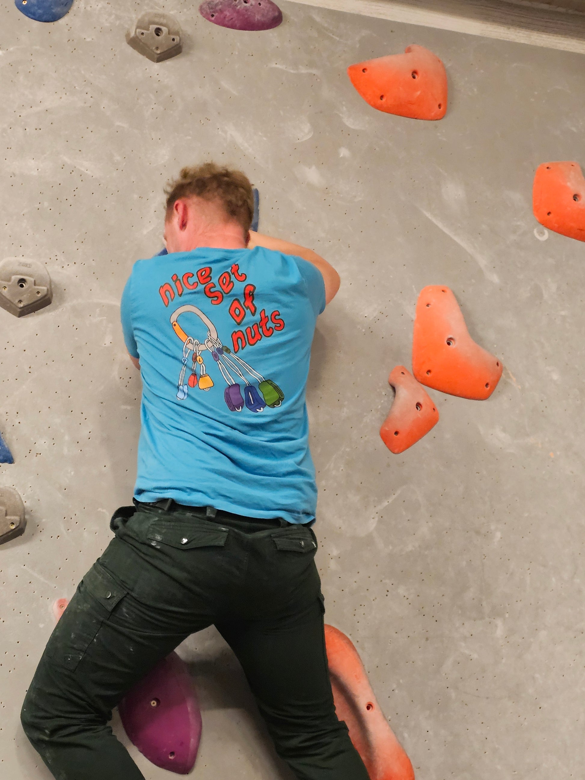 Person on a bouldering wall wearing the "Nice Set Of Nuts" T-Shirt in light blue. 